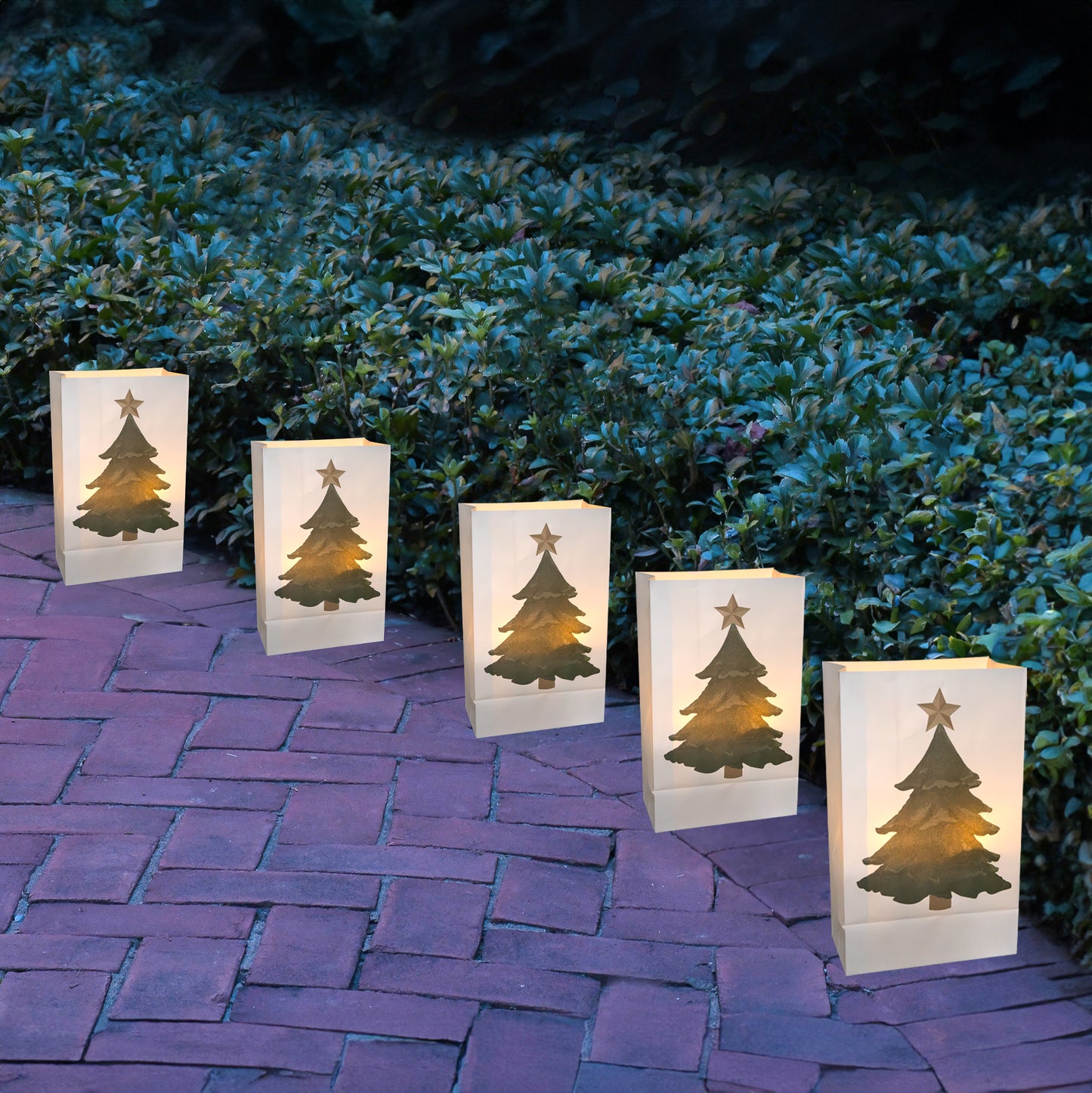 Five glowing white paper luminaria bags with a printed design of a Christmas tree and a star at the top lined along a brick walkway.