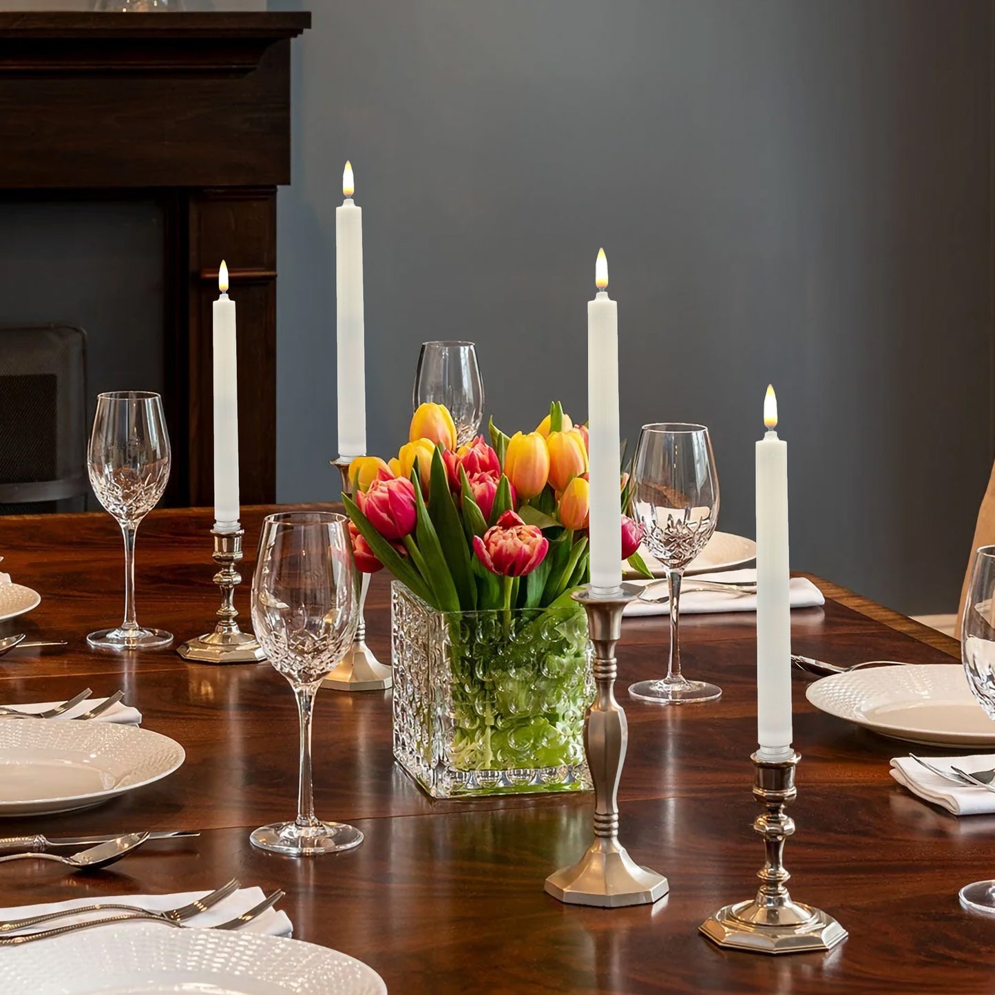 A set of four battery operated 3D wick flame taper candles with visible wicks and amber-colored flames with a vase of flowers and wine glasses at the place settings.