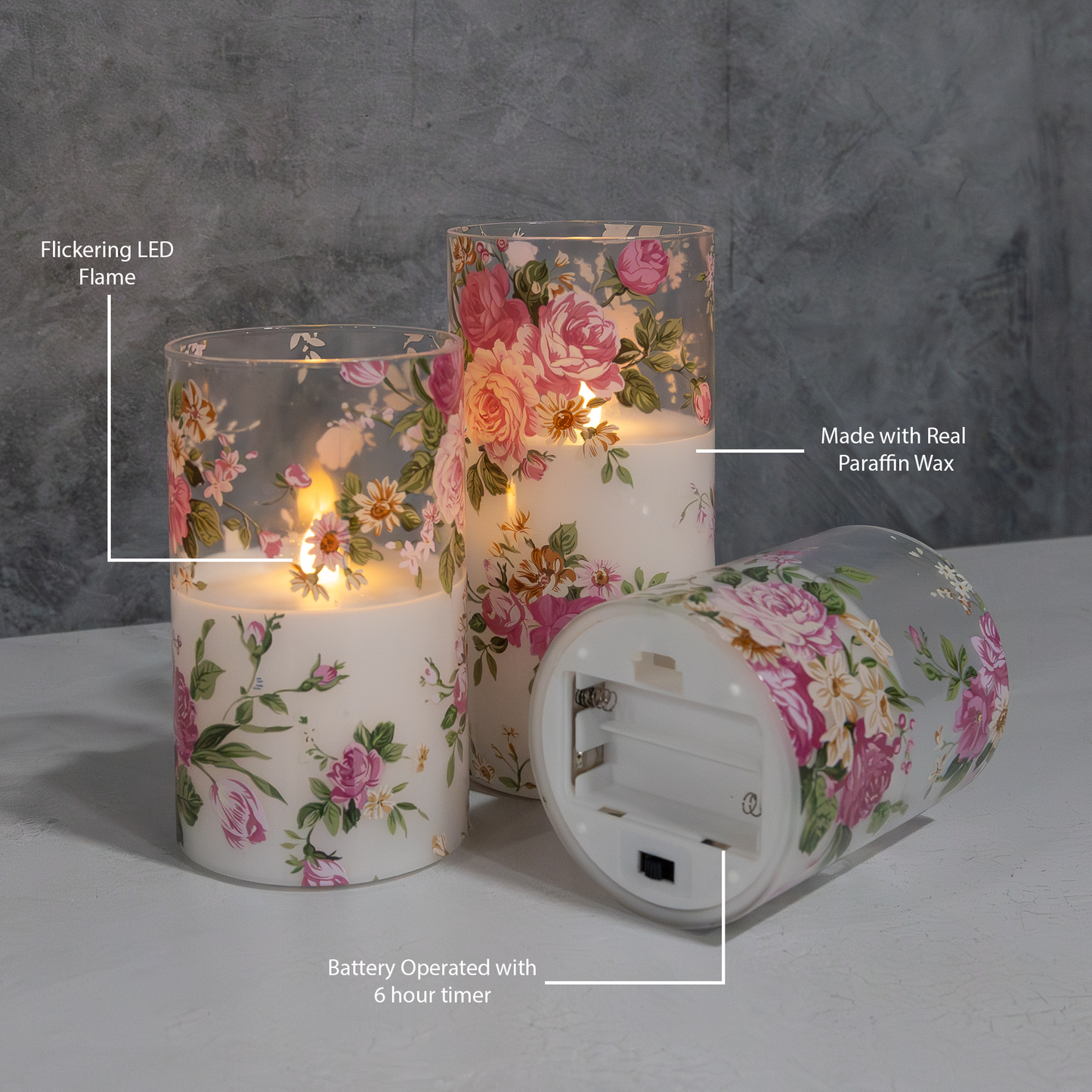 Set of three battery operated clear glass hurricane candles with a floral design, featuring a wax candle inside and an LED light visible through the glass showing battery placement.