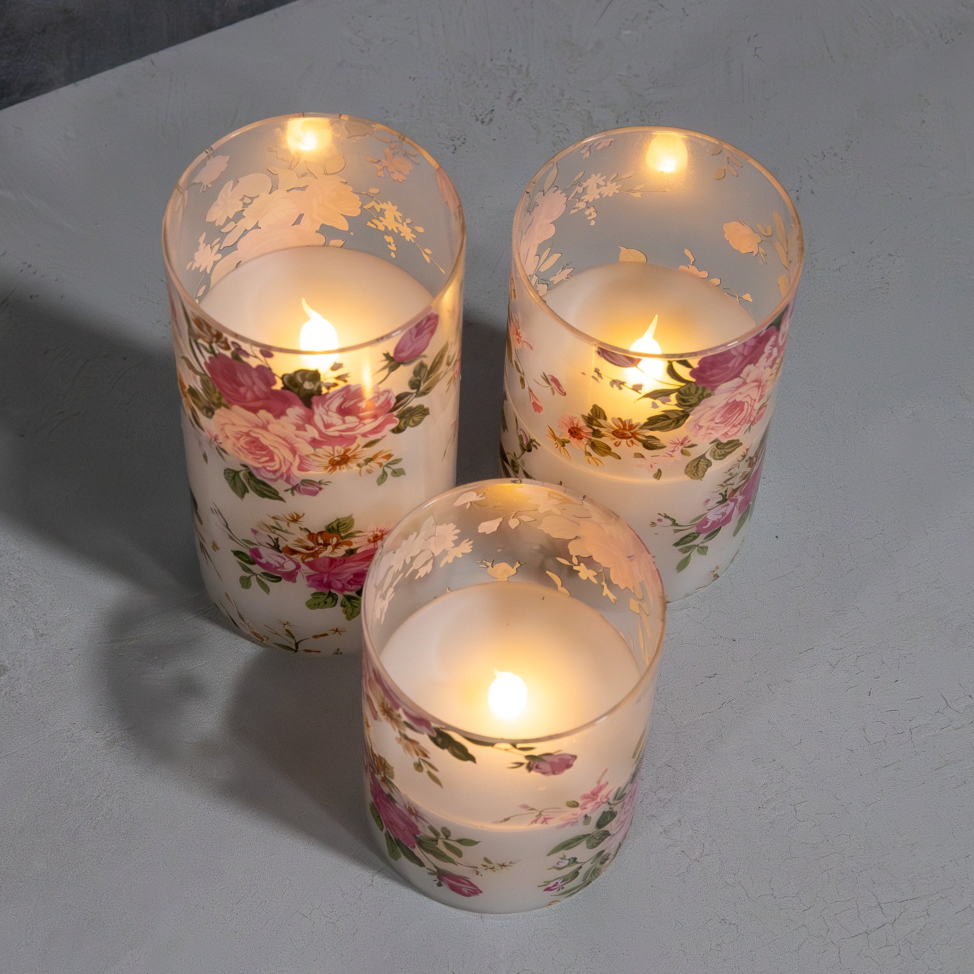 Set of three battery operated clear glass hurricane candles with a floral design, featuring a wax candle inside and an LED light visible through the glass.