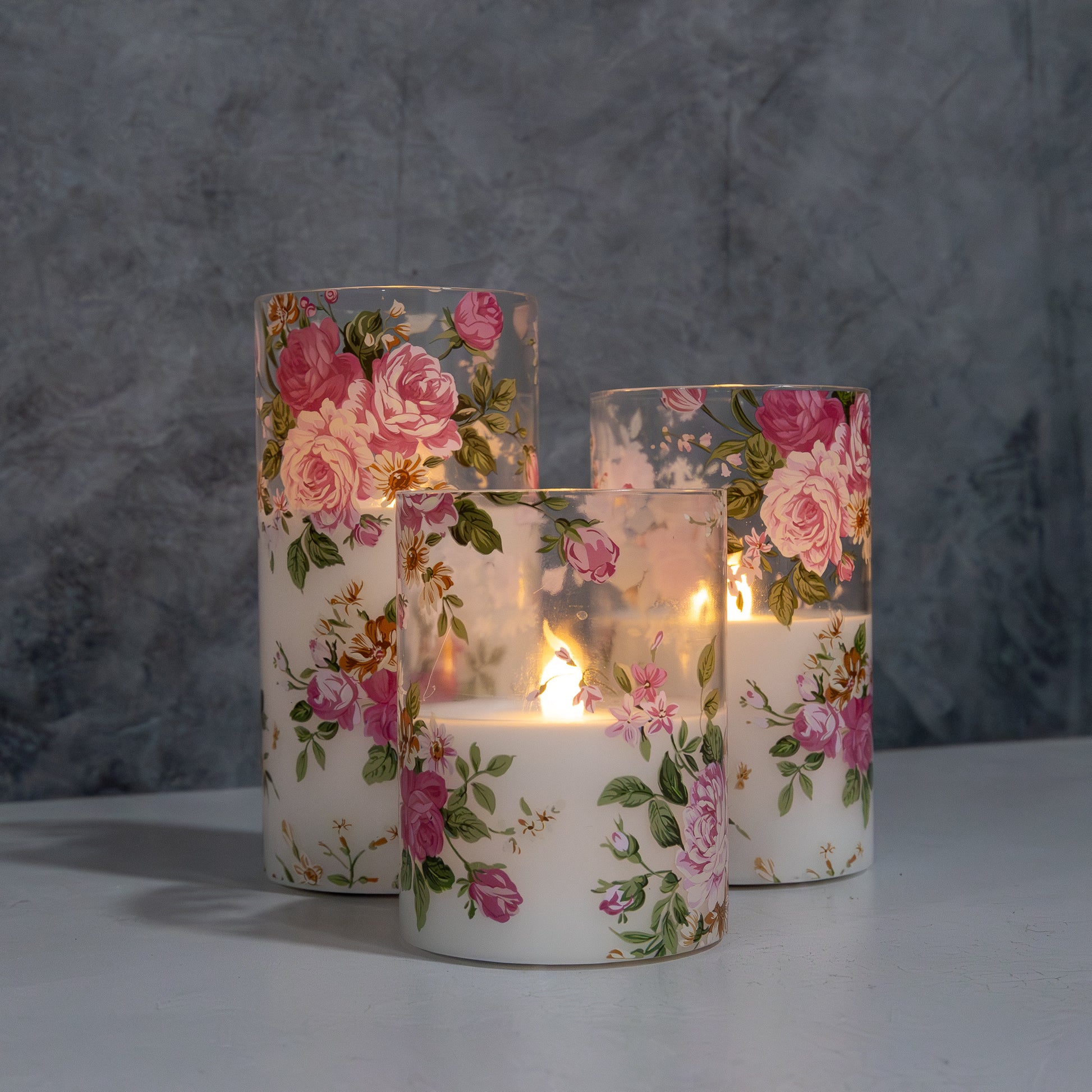 Set of three battery operated clear glass hurricane candles with a floral design, featuring a wax candle inside and an LED light visible through the glass.