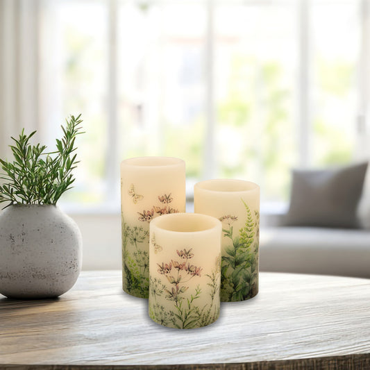 A set of three white wax candles with floral designs and greenery motifs, featuring an amber flickering LED light on a table with a plant.