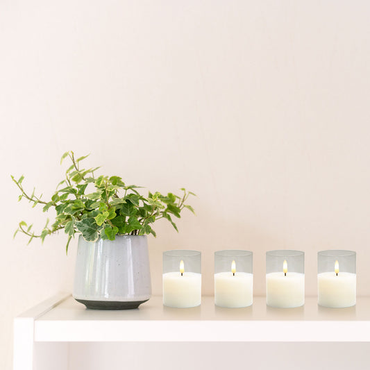 A set of four clear, battery-operated 3D wick flame LED candles with a soft white glow along with a plant displayed on a white shelf.