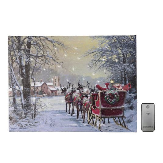 A canvas print depicting Santa Claus riding in a sleigh pulled by reindeer, with a snowy town in the background. The canvas is lighted with twinkling cool white LED lights, and there's a remote control next to it.