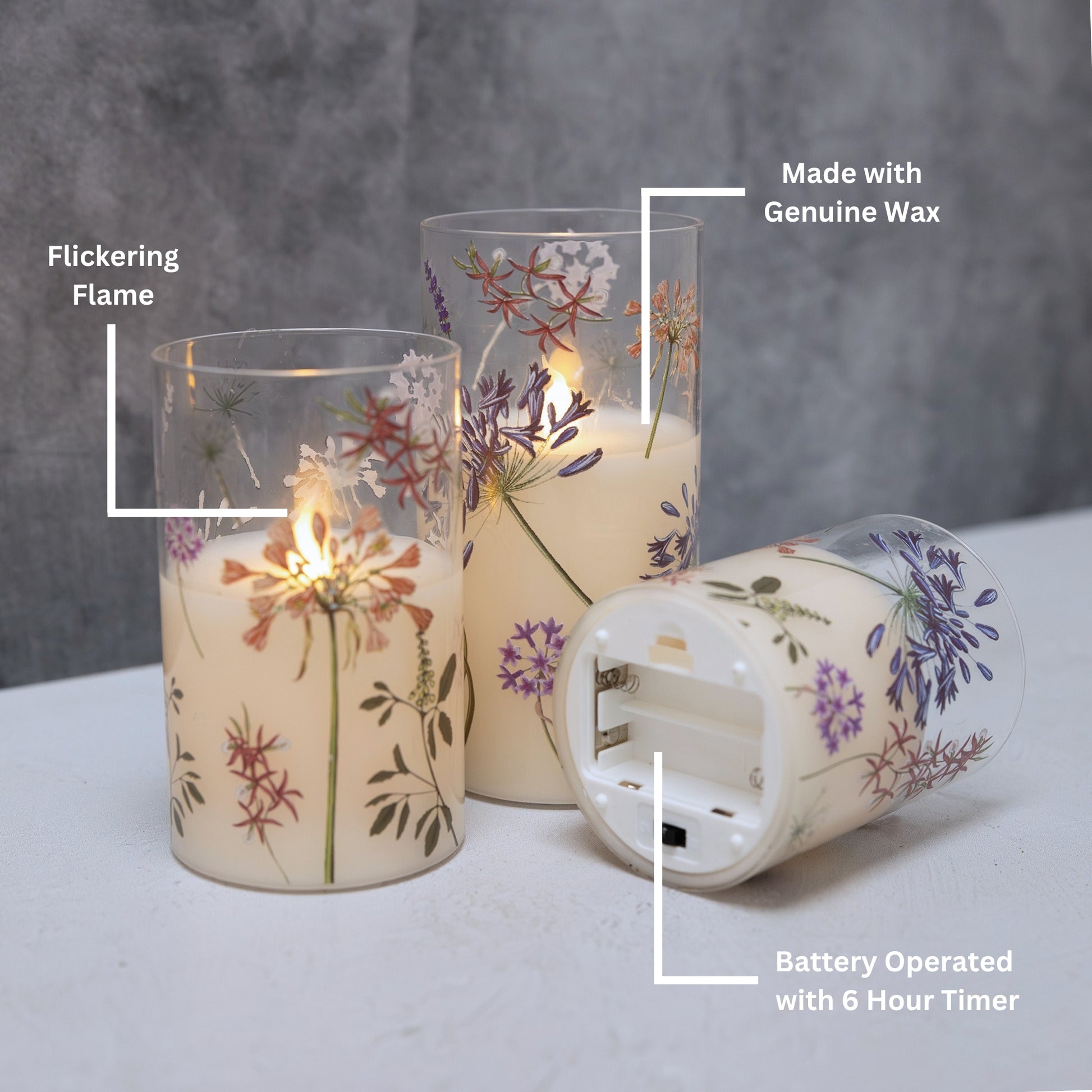 Set of three battery operated clear glass hurricane candles with a floral design, featuring a wax candle inside and an LED light visible through the glass showing battery placement.