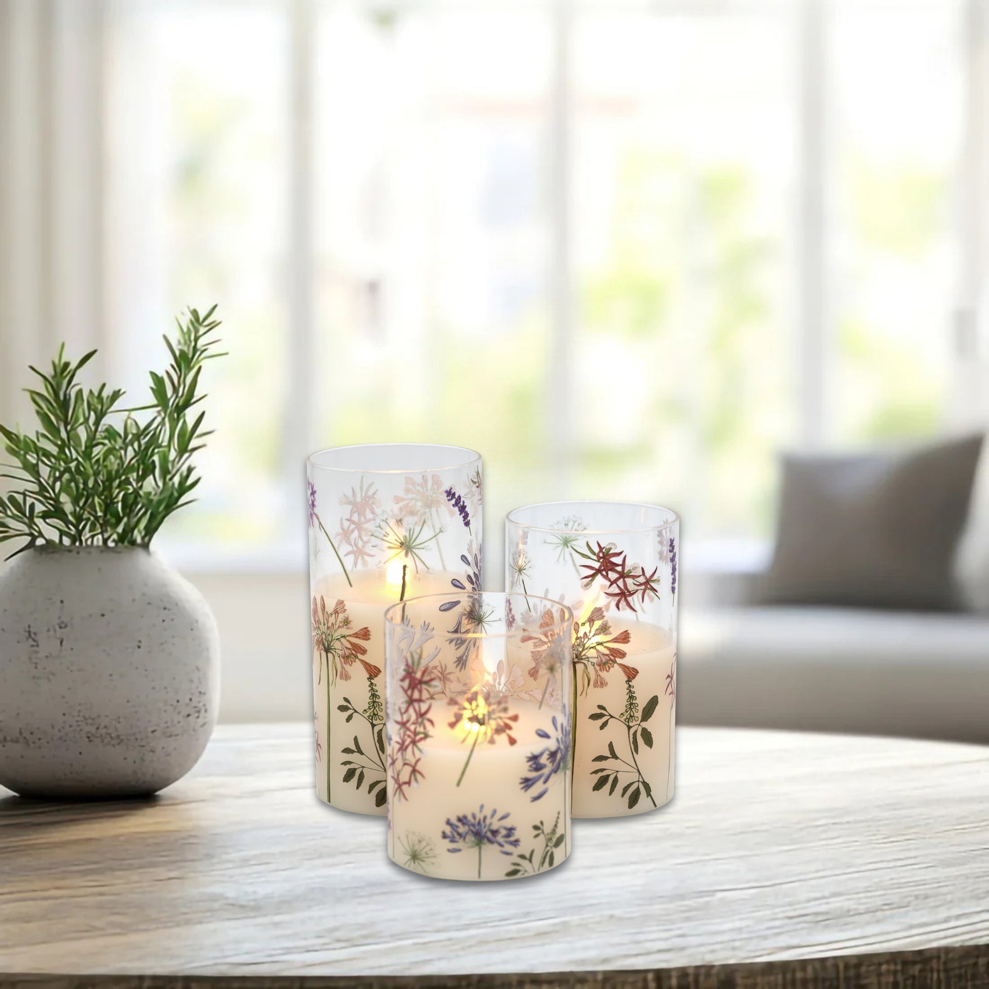 Set of three battery operated clear glass hurricane candles with a floral design, featuring a wax candle inside and an LED light visible through the glass on a table with a plant.